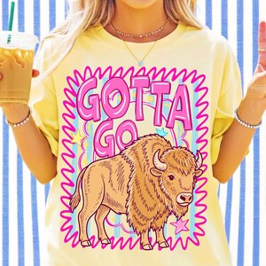 May include: A pale yellow t-shirt featuring a graphic print of a brown bison and the text "GOTTA GO" in large pink letters. The design is framed by a jagged pink border and includes small yellow and pink stars against a light blue striped background.