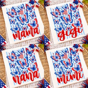 May include: A white t-shirt featuring a patriotic design with red, white, and blue butterflies, American flag motifs, and light blue vertical stripes. The shirts display the red cursive text mama, gigi, nana, or mimi across the lower section.