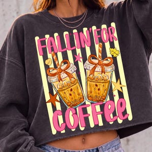 May include: A charcoal gray long-sleeve sweatshirt featuring a graphic print of two iced coffee cups with orange bows, surrounded by yellow vertical stripes, stars, and leaves. The text "FALLIN' FOR COFFEE" is printed in bright pink lettering.