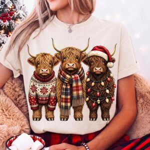 May include: A cream-colored t-shirt featuring a graphic of three brown Highland cows. The left cow wears a red and white snowflake sweater, the middle cow wears a plaid scarf, and the right cow wears a red Santa hat and string lights.