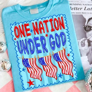 May include: A light blue t-shirt featuring the red and blue text ONE NATION UNDER GOD. Below the text, three American flags hang from a clothesline. The design is framed by a scalloped blue border with small red and blue stars scattered around.