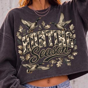 May include: A charcoal gray cropped sweatshirt featuring a graphic print of three flying mallard ducks surrounded by oak leaves. The text "HUNTING SEASON" is printed in the center with a camouflage pattern fill and a dark outline.