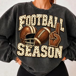 Puede incluir: Una sudadera de cuello redondo gris carb&oacute;n con un estampado gr&aacute;fico que incluye el texto FOOTBALL SEASON en una fuente de color crema con lunares. El dise&ntilde;o muestra un casco de f&uacute;tbol americano, un bal&oacute;n de f&uacute;tbol y un dedo de espuma marr&oacute;n.