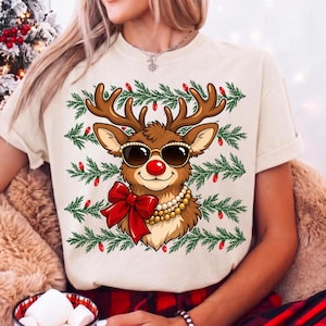 Puede incluir: Una camiseta de color crema con el gr&aacute;fico de un reno que lleva gafas de sol negras, un collar de perlas y un gran lazo rojo. El reno est&aacute; rodeado por filas horizontales de ramas de pino verdes decoradas con peque&ntilde;as luces rojas.