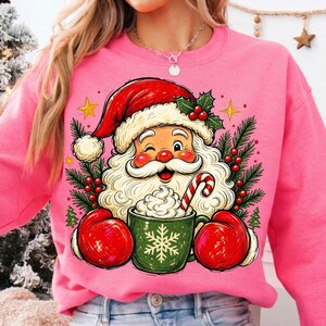 May include: A bright pink sweatshirt featuring a graphic of Santa Claus wearing red mittens and a red hat, winking while holding a green mug filled with whipped cream and a candy cane, surrounded by pine branches, holly berries, and gold stars.