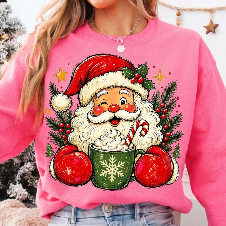 May include: A bright pink sweatshirt featuring a graphic of Santa Claus wearing red mittens and a red hat, winking while holding a green mug filled with whipped cream and a candy cane, surrounded by pine branches, holly berries, and gold stars.