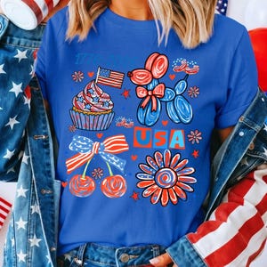 May include: A royal blue t-shirt featuring a graphic print with the text 1776-2026 and USA. The design includes a cupcake with an American flag, a balloon dog, cherries with a striped bow, and a flower, all in red, white, and blue patriotic colours.
