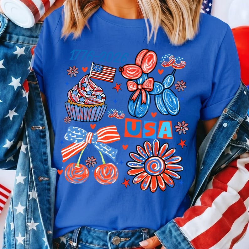 May include: A royal blue t-shirt featuring a graphic print with the text 1776-2026 and USA. The design includes a cupcake with an American flag, a balloon dog, cherries with a striped bow, and a flower, all in red, white, and blue patriotic colours.