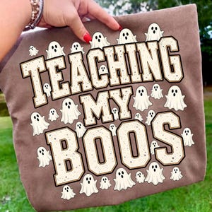 May include: A brown fabric item featuring a graphic print of numerous small white ghosts and the text "TEACHING MY BOOS" in large, block-style letters with a speckled, light-colored fill and dark outlines, arranged in a three-line layout.
