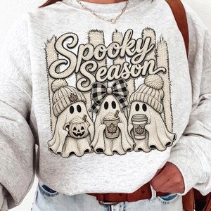 May include: A light gray crewneck sweatshirt featuring a graphic of three ghosts wearing knit beanies. The ghosts hold a pumpkin mug, a cup with a maple leaf, and a coffee cup. The text Spooky Season is printed above in a bold, cursive font.