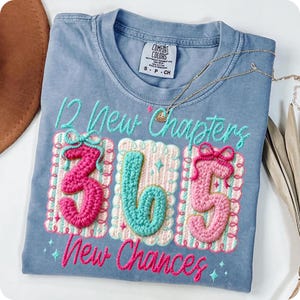 Puede incluir: Camiseta azul claro Comfort Colors con el texto bordado "12 New Chapters 365 New Chances". Los n&uacute;meros son de estilo crochet, con detalles rosas, turquesas y blancos. Un collar dorado descansa sobre la camiseta.