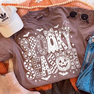 May include: A brown short-sleeved t-shirt featuring a white graphic print that reads SPOOKY SEASON. The design includes a ghost with a bow, a jack-o'-lantern with a bow, bats, and stars. The letters are filled with various patterns like checkers, dots, and flowers.