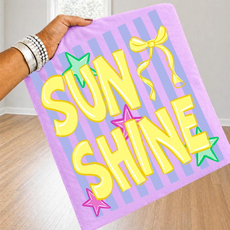 May include: A rectangular, lavender blanket with the words "SUN SHINE" in large, yellow, bubble letters. The blanket has blue vertical stripes and star and bow accents. The blanket is held by a person.