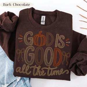 May include: A dark chocolate brown crewneck sweatshirt featuring the text "GOD IS GOOD all the time" in a multi-line, retro-style font. The letter "O" in "GOD" is replaced by an orange pumpkin graphic, and two line-art bows flank the text.