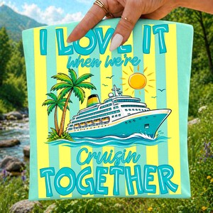 K&ouml;nnte beinhalten: Ein hellblau und gelb gestreifter Stoff mit einer Grafik eines Kreuzfahrtschiffes, zwei Palmen und einer gelben Sonne. Der Text lautet I LOVE IT when we're Cruisin TOGETHER in blauen Buchstaben. Das Design zeigt blaue Meereswellen.