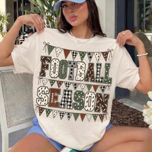 May include: A cream-colored short-sleeve t-shirt featuring a graphic print of triangular pennant banners and the text "FOOTBALL SEASON" in large block letters. The design incorporates various patterns including plaid, footballs, and stars.