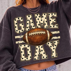 Op de afbeelding: Een antracietgrijs sweatshirt met lange mouwen en een grafische print met de tekst GAME DAY in grote, cr&egrave;mekleurige blokletters met zwarte stippen. Een bruine American football met witte veters staat centraal tussen de twee woorden.