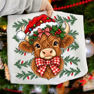 Puede incluir: Una camiseta blanca con una ilustraci&oacute;n gr&aacute;fica de una vaca Highland marr&oacute;n que lleva un gorro de Pap&aacute; Noel rojo con luces navide&ntilde;as, una corona de acebo verde con bayas rojas y un lazo de cuadros rojo y blanco bajo la barbilla.