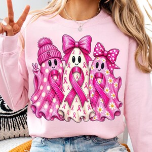 Puede incluir: Una sudadera rosa claro con un gr&aacute;fico de tres fantasmas. El fantasma de la izquierda lleva un gorro rosa y hace el signo de la paz. El fantasma central tiene un gran lazo rosa. El de la derecha tiene un lazo rosa de lunares y margaritas.