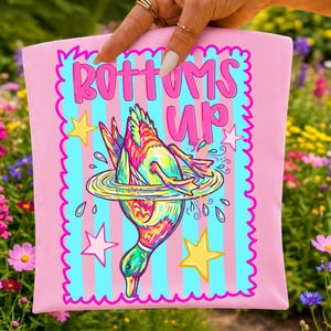 May include: A light pink fabric item featuring a graphic of a colorful duck diving into water with the text Bottoms Up. The design includes a light blue and pink striped background, a scalloped border, and yellow and white stars surrounding the duck.