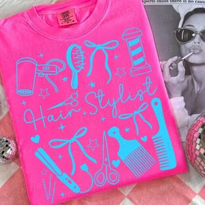 May include: A bright pink t-shirt with a light blue design featuring hair styling tools and the words "Hair Stylist". The design includes a hair dryer, comb, scissors, and a barber pole. The shirt has a crew neck.