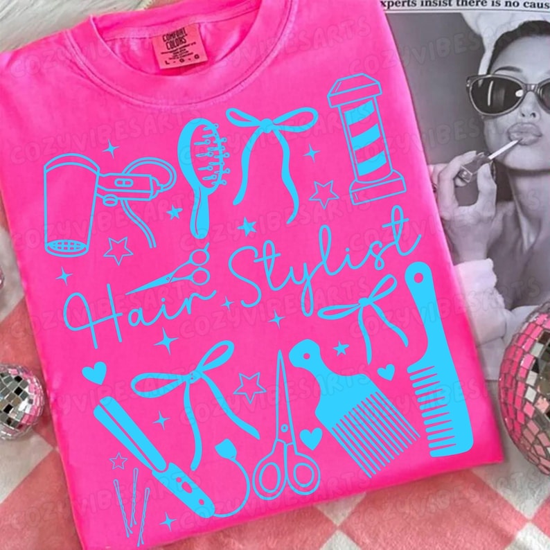 May include: A bright pink t-shirt with a light blue design featuring hair styling tools and the words "Hair Stylist". The design includes a hair dryer, comb, scissors, and a barber pole. The shirt has a crew neck.
