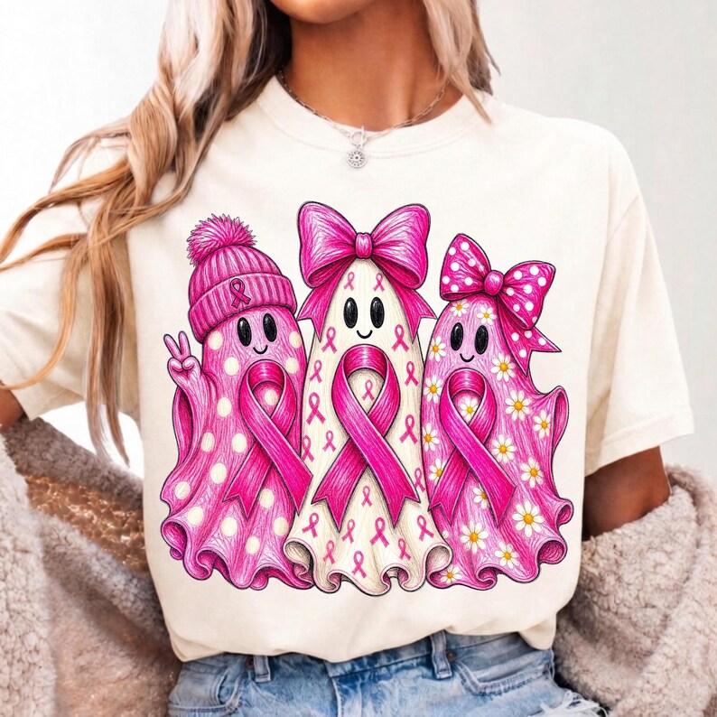 Puede incluir: Una camiseta de color crema con un gr&aacute;fico de tres fantasmas. El fantasma de la izquierda lleva un gorro rosa y hace el signo de la paz. El fantasma central tiene un lazo rosa grande y cintas de concienciaci&oacute;n sobre el c&aacute;ncer de mama.