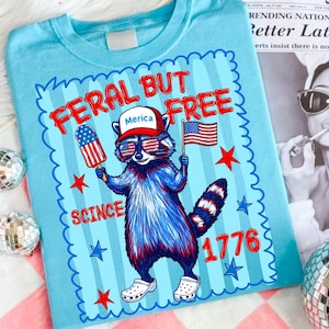 May include: A light blue t-shirt featuring a graphic of a raccoon wearing a red hat with the text Merica, sunglasses with American flag patterns, and white clogs. The raccoon holds a flag and a popsicle. Text reads FERAL BUT FREE SCINCE 1776.