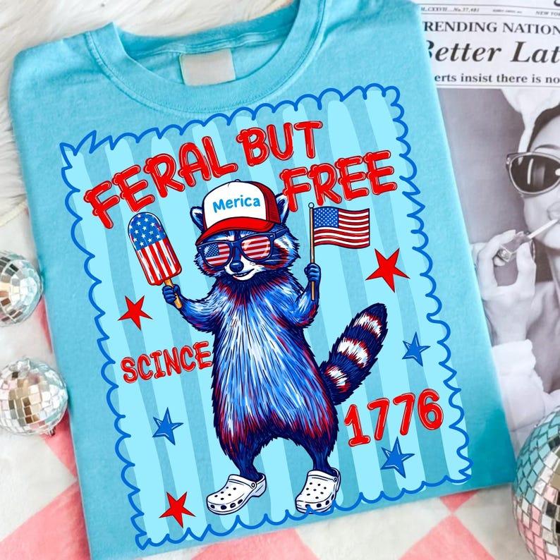 May include: A light blue t-shirt featuring a graphic of a raccoon wearing a red hat with the text Merica, sunglasses with American flag patterns, and white clogs. The raccoon holds a flag and a popsicle. Text reads FERAL BUT FREE SCINCE 1776.