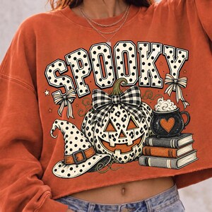 May include: An orange long-sleeve sweatshirt featuring a graphic print with the text SPOOKY in white polka-dot letters. The design includes a leopard-print pumpkin with a plaid bow, a polka-dot witch hat, a black mug with a heart, and three books.