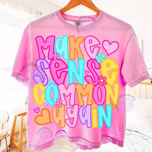 May include: Pink t-shirt with the phrase "Make Sense Common Again" in colorful, bubble-style letters. The design includes hearts and flowers. The shirt is hanging on a wooden hanger.