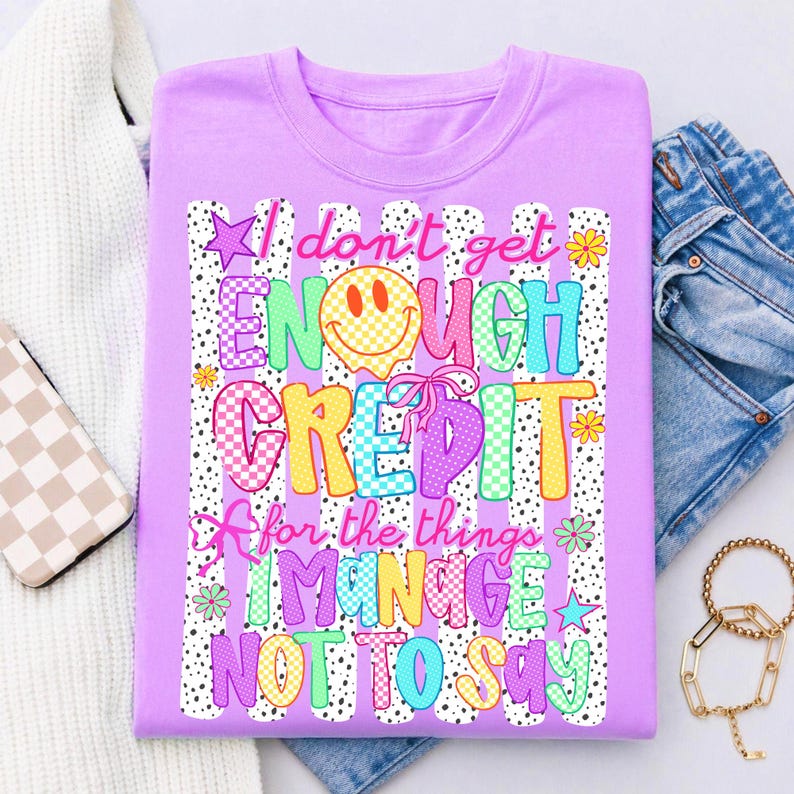 Pode incluir: Uma t-shirt roxo-claro com um gr&aacute;fico que inclui o texto I don't get ENOUGH CREDIT for the things I MANAGE NOT TO SAY. O design apresenta uma cara sorridente amarela, la&ccedil;os cor-de-rosa, estrelas, flores e riscas verticais com pontos pretos.