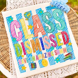 May include: A white t-shirt featuring a blue striped background with the text "CLASS DISMISSED" and "HAVE AN AMAZING SUMMER" in colorful, scribbled-style letters. The design includes illustrations of a globe, stars, flowers, pencils, and crayons.