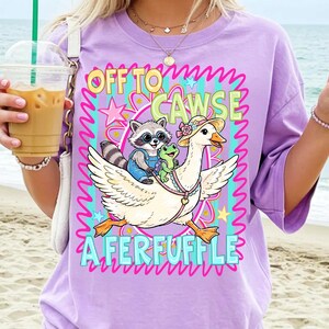 May include: A lilac-colored t-shirt features a whimsical graphic of a goose wearing a hat, carrying a raccoon and a frog. The text reads "OFF TO CAWSE A FERFUFFLE" in colorful, playful lettering. The design includes stars and wavy lines.