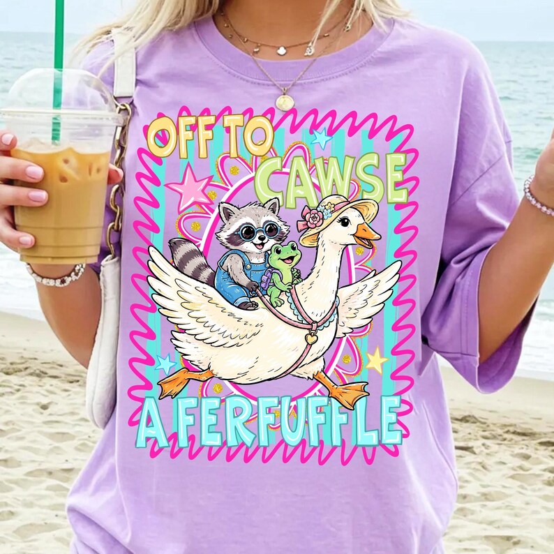 May include: A lilac-colored t-shirt features a whimsical graphic of a goose wearing a hat, carrying a raccoon and a frog. The text reads "OFF TO CAWSE A FERFUFFLE" in colorful, playful lettering. The design includes stars and wavy lines.