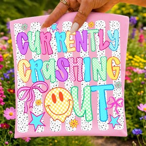 Pu&ograve; includere: Una pochette rettangolare rosa chiaro con un motivo a righe a pois bianchi e neri. Il testo "CURRENTLY CRASHING OUT" &egrave; stampato in lettere colorate, a scacchi e a tinta unita. Il design include una faccina sorridente gialla e fiocchi rosa.