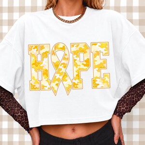 May include: A white short-sleeved t-shirt featuring the word HOPE in large block letters with a yellow and white camouflage pattern. The letter O is replaced by a yellow awareness ribbon. The shirt is worn over a brown leopard print long-sleeved top.