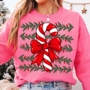 May include: A bright pink crewneck sweatshirt featuring a central graphic of a large red and white striped candy cane tied with a prominent red bow. The background of the design consists of horizontal rows of green pine branches and red lights.