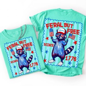 May include: A light green t-shirt featuring a graphic of a raccoon wearing a red, white, and blue hat with the text Merica, sunglasses, and white clogs. The raccoon holds a flag and a popsicle. Text reads FERAL BUT FREE SCINCE 1776.