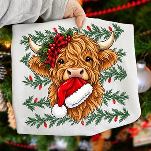 May include: A white t-shirt featuring a graphic illustration of a fluffy brown Highland cow with a red and black buffalo plaid bow on its head, holding a red and white Santa hat in its mouth, surrounded by green pine branches and red lights.