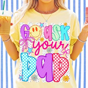 Op de afbeelding: Een lichtgeel t-shirt met een grafische print met de tekst "GO ASK YOUR DAD". Het ontwerp bevat een geel smileygezicht, roze en blauwe ruitpatronen, kleine sterretjes, bloemen en strikjes tegen een achtergrond van zwarte stippen.
