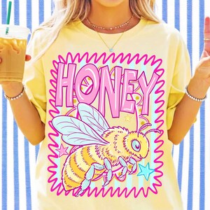May include: A pale yellow t-shirt featuring a graphic print with the word HONEY in large pink letters. Below the text is a detailed illustration of a yellow and black bee with light blue wings, surrounded by a pink squiggly border and stars.