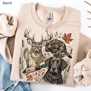 May include: A sand-colored crewneck sweatshirt featuring a graphic print of a deer, a mallard duck, a camouflage baseball cap, and a black Labrador retriever. The design includes cattails and autumn leaves surrounding the wildlife imagery.