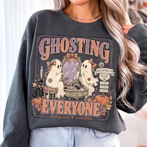 May include: A dark gray crewneck sweatshirt featuring a graphic print with the text GHOSTING EVERYONE. The design shows two ghosts with bows, a black cat, a vanity mirror, pumpkins, and a digital display reading SOCIAL BATTERY: 0%.