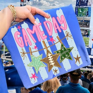 May include: A blue fabric pouch featuring a vertical striped pattern and the pink text GAME DAY. A clothesline graphic displays three star-shaped ornaments: a light green play diagram star, a brown football star, and a green football field star.