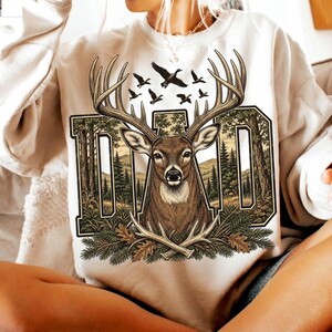 May include: A cream-coloured crewneck sweatshirt featuring a large graphic print. The design displays the word DAD in block letters filled with a forest scene, a central deer head with large antlers, flying ducks, and crossed antlers at the base.
