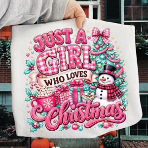 May include: A white t-shirt featuring a colorful Christmas graphic with the text "JUST A GIRL WHO LOVES CHRISTMAS". The design includes a decorated pine tree, a snowman with a plaid scarf, wrapped gifts, and a mug of hot cocoa with whipped cream.