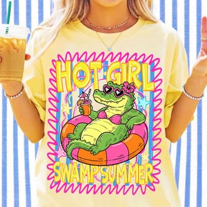 K&ouml;nnte beinhalten: Ein blassgelbes T-Shirt mit einer Grafik eines gr&uuml;nen Alligators, der eine herzf&ouml;rmige Sonnenbrille und ein rosa Bikini-Oberteil tr&auml;gt, w&auml;hrend er in einem orange-rosa Schwimmreifen treibt, mit dem Text "HOT GIRL SWAMP SUMMER" in gelben Buchstaben.