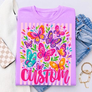 May include: A light purple t-shirt featuring a graphic print of colourful butterflies in shades of pink, orange, yellow, and teal against a background of vertical light purple stripes. The word CUSTOM is printed in a bold, cursive pink font at the bottom.