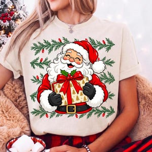 May include: A cream-colored t-shirt featuring a graphic of Santa Claus with a white beard and red hat, holding a gold gift box with a red bow. The design is framed by green pine branches and small red Christmas lights in a repeating pattern.